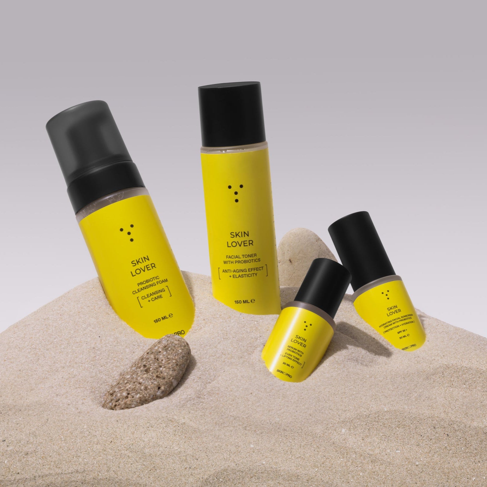 Four yellow skincare bottles with black caps on a sandy surface